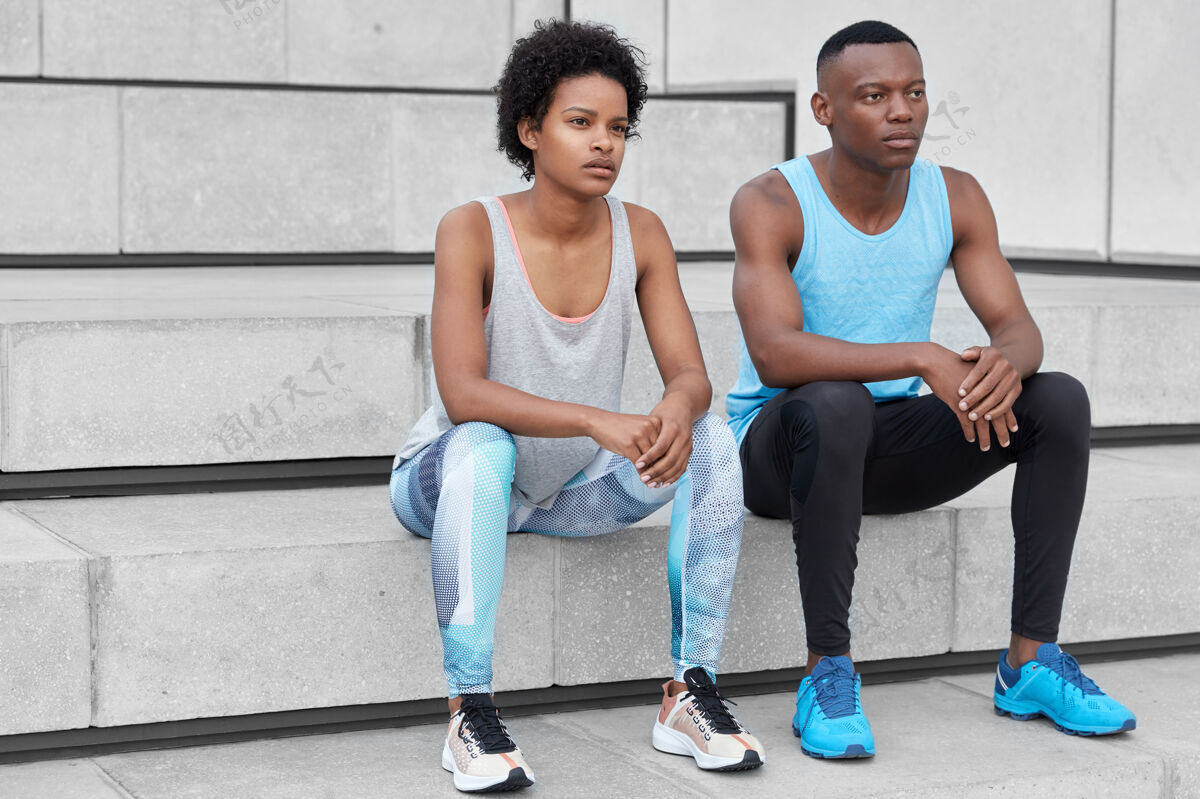 男孩一对皮肤黝黑的情侣穿着运动服 穿着舒适的运动鞋 有氧训练后休息 坐在城市楼梯旁 专注于远处 有点累男女朋友都喜欢运动女人非洲男人