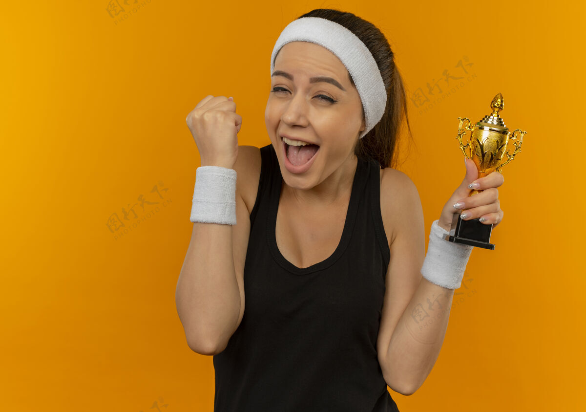拳头身穿运动服 头箍紧握拳头 手持奖杯的年轻健身女士站在橙色的墙上 快乐而兴奋抱着运动人