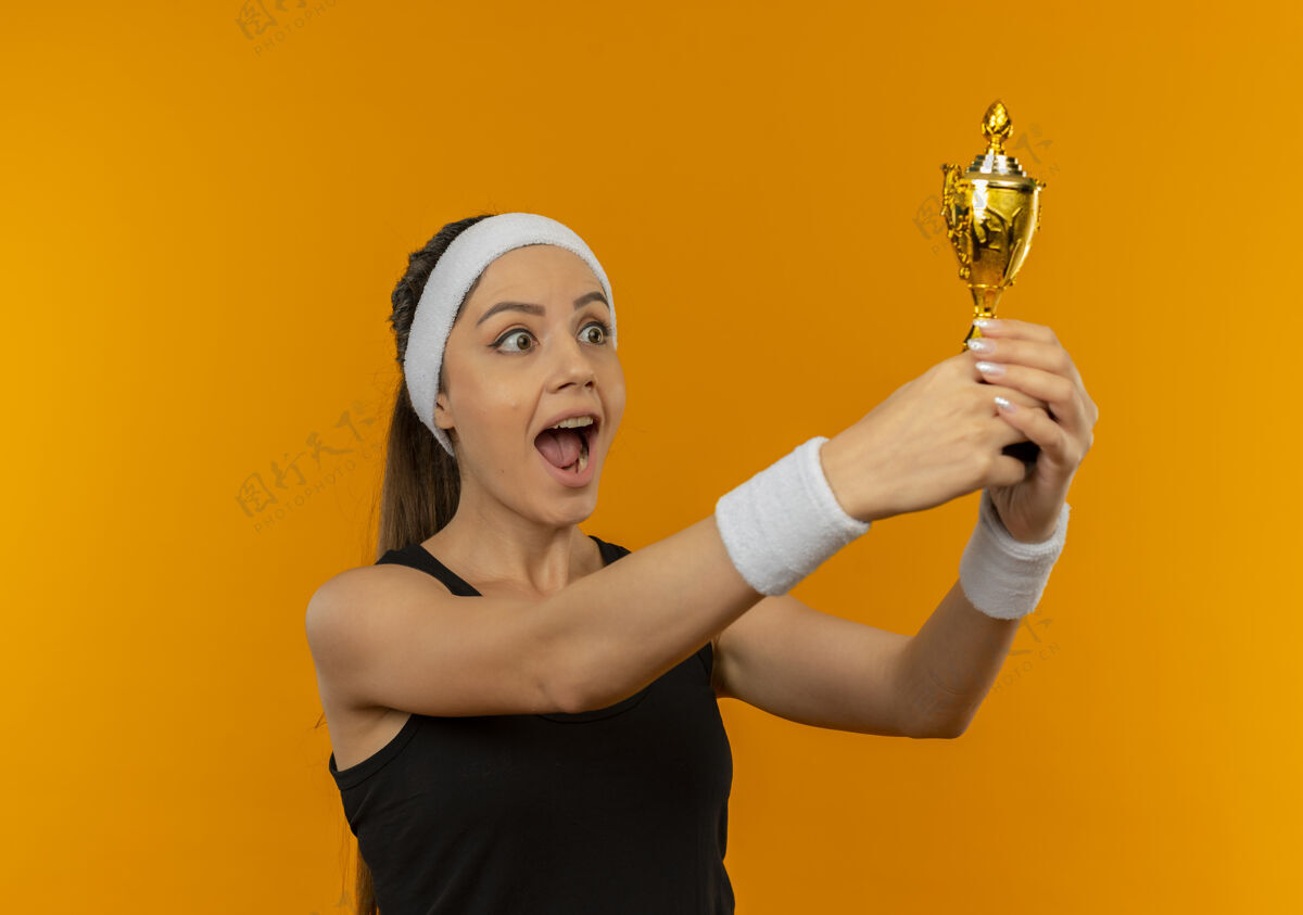 姿势身穿运动服 头箍 手持奖杯的年轻健身女士站在橙色的墙上 快乐而兴奋兴奋女人快乐