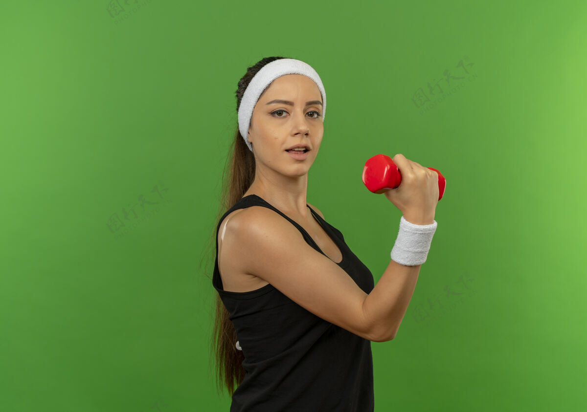 抱身穿运动服 头箍 举着哑铃的年轻健身女士站在绿色的墙壁上 看起来很自信健康锻炼看