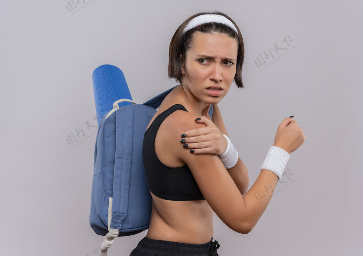 运动穿着运动服 背着背包和瑜伽垫的年轻健身女士 看起来不舒服 摸着肩膀 站在白墙上感觉疼痛看姿势运动装