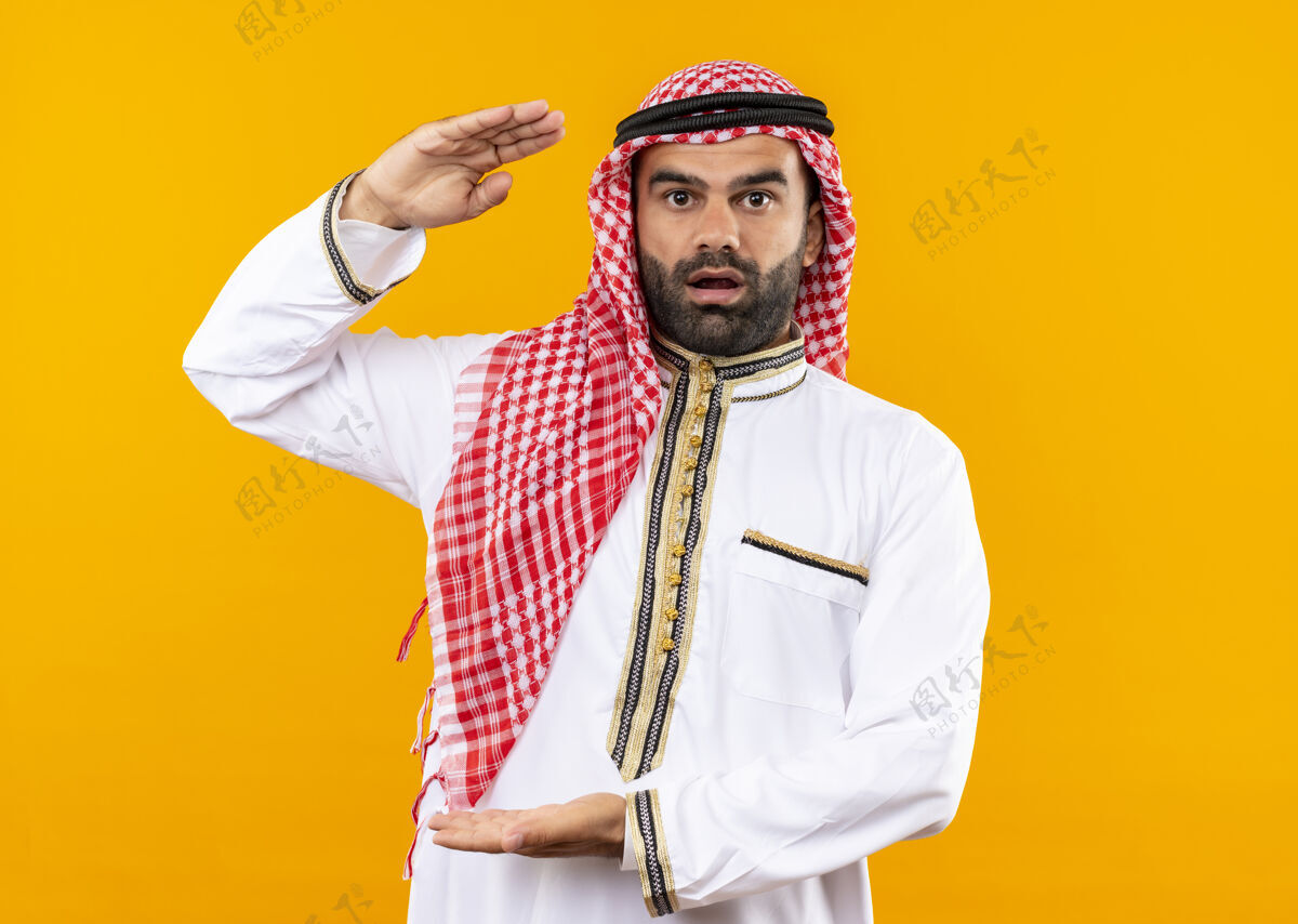 显示身着传统服装的阿拉伯商人看起来很惊讶地展示着大尺寸的手势 站在橙色的墙上测量符号手势阿拉伯语大小