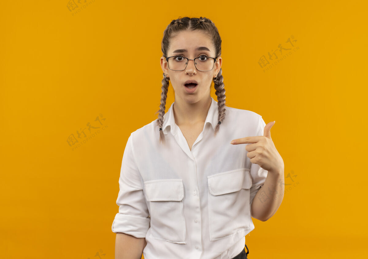 衬衫戴着眼镜 梳着辫子 穿着白衬衫的年轻女学生困惑地指着站在橘色墙上的自己看着前方辫子手势人