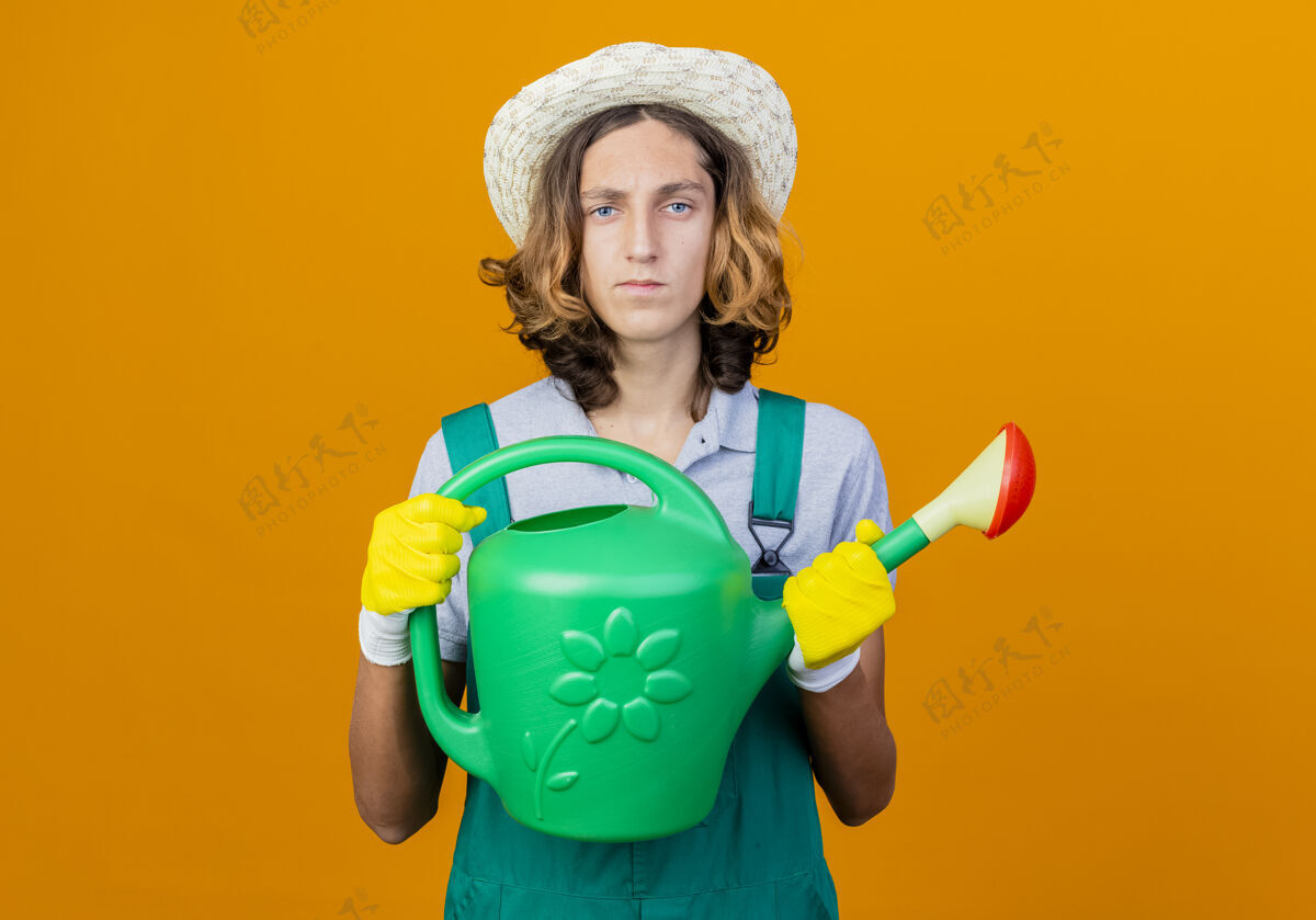 帽子年轻的园丁戴着橡胶手套 穿着连体衣 戴着帽子 手里拿着浇水罐连身衣手套抱