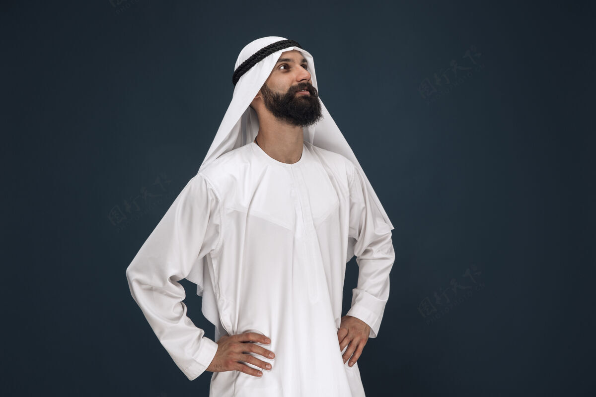 手势深蓝色工作室背景上的半身阿拉伯沙特商人肖像年轻男模站着微笑商业 金融 面部表情 人类情感的概念成人胡须阿拉伯语