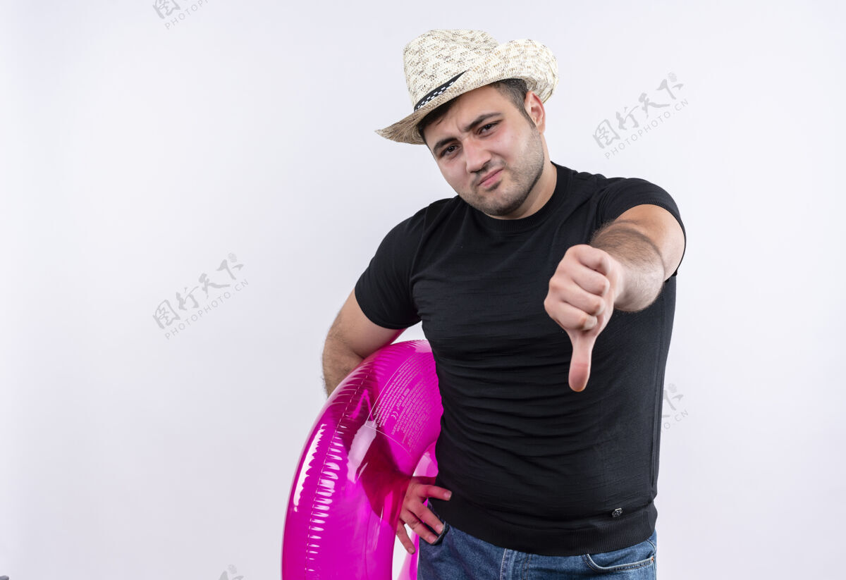 站着不高兴的年轻旅行家男子身穿黑色t恤 戴着夏季帽子 手持充气戒指 大拇指朝下 站在白色的墙上拇指夏天展示