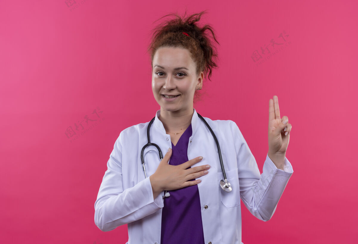 微笑身穿白大褂 手持听诊器 微笑着站在粉色墙壁上宣誓的年轻女医生宣誓年轻人听诊器