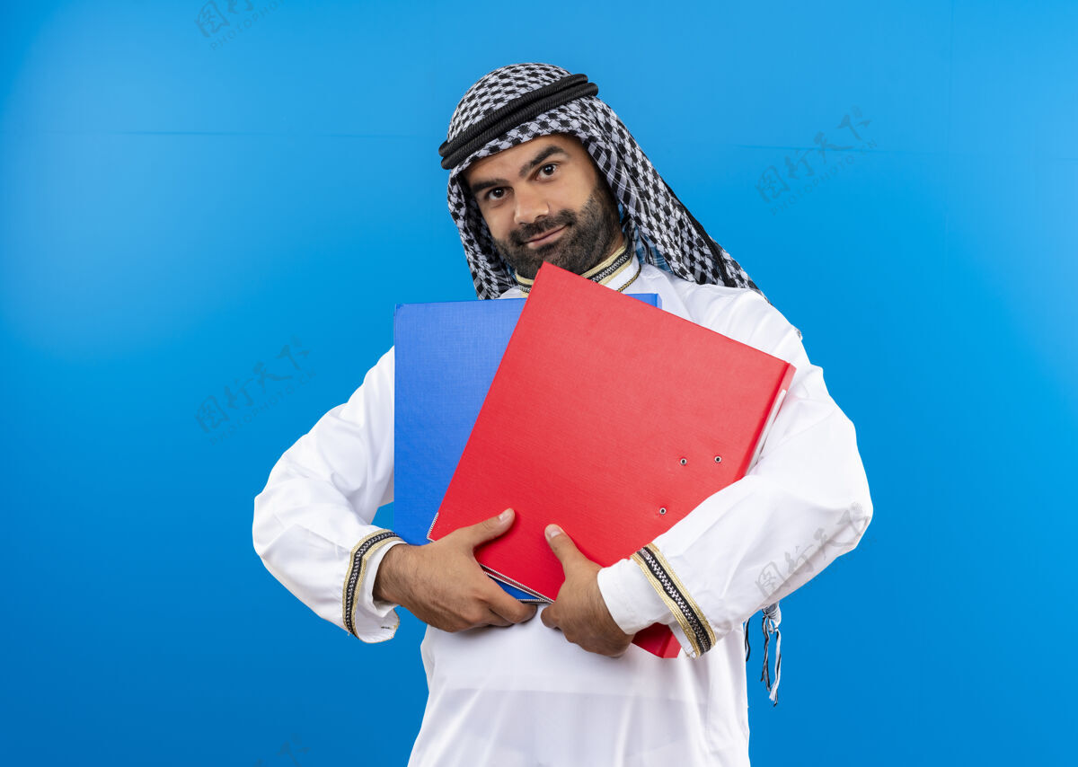 脸身着传统服装的阿拉伯商人手拿两个文件夹 脸上带着微笑站在蓝色的墙上站阿拉伯语文件夹