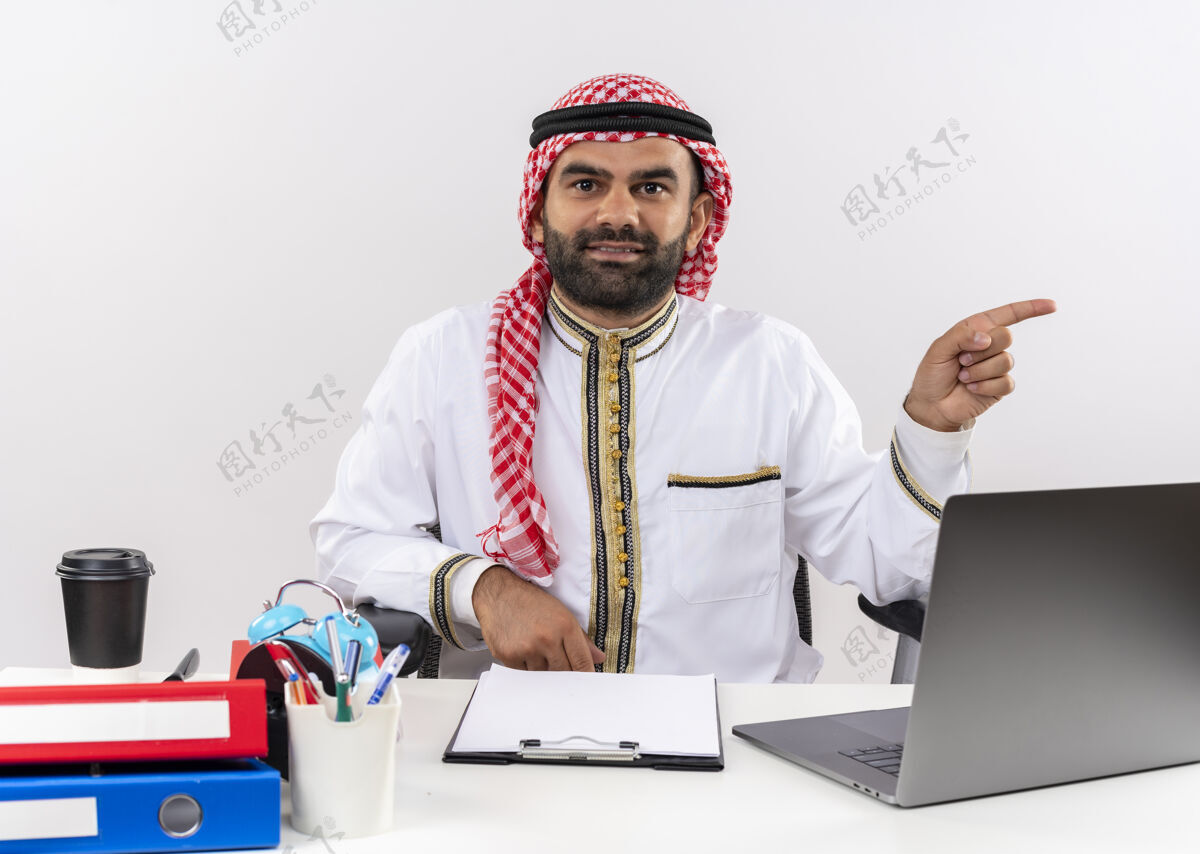 手指身着传统服装的阿拉伯商人正手拿笔记本电脑 手指指向一边 自信地坐在办公室的桌子旁指阿拉伯语穿