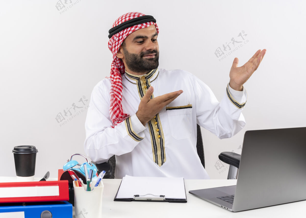 信心身着传统服装的阿拉伯商人坐在桌旁 手持笔记本电脑 双臂指向一边 看上去自信地在办公室工作阿拉伯语传统工作