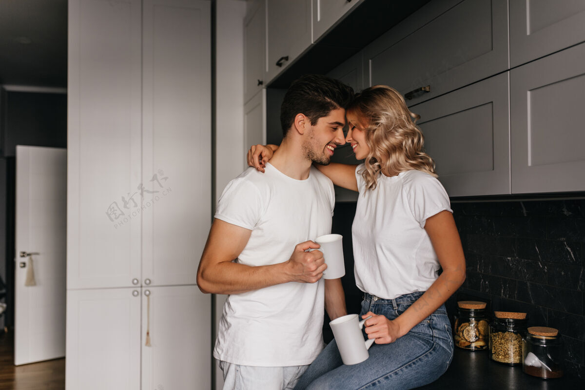 室内迷人的卷发女孩在早餐时拥抱丈夫帅哥拿着咖啡在厨房摆姿势的室内肖像午餐积极放松