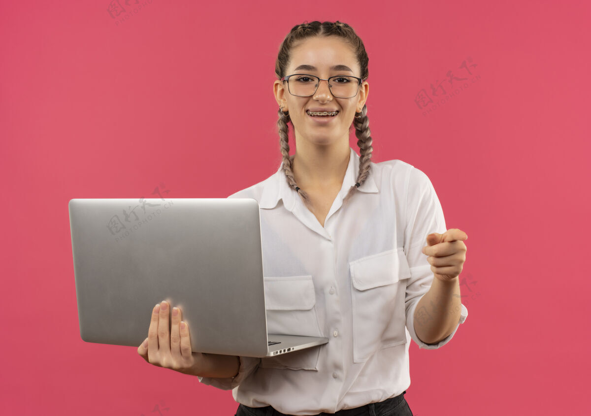市民戴着眼镜 梳着辫子 穿着白衬衫的年轻女学生手里拿着笔记本电脑 手指指着前面 站在粉色的墙上欢快地笑着辫子眼镜女孩