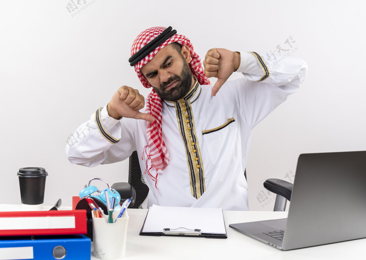 拇指身着传统服装的阿拉伯商人坐在桌子旁 手里拿着笔记本电脑 看起来很不高兴 在办公室工作时大拇指朝下看笔记本电脑传统