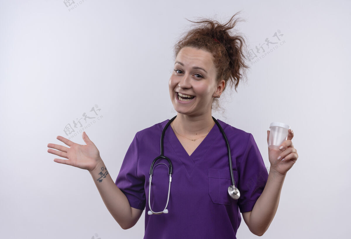 微笑年轻的女医生穿着医疗制服 手持听诊器 举着测试罐 微笑着 愉快地抬起手臂站在白色的墙上胳膊罐子制服