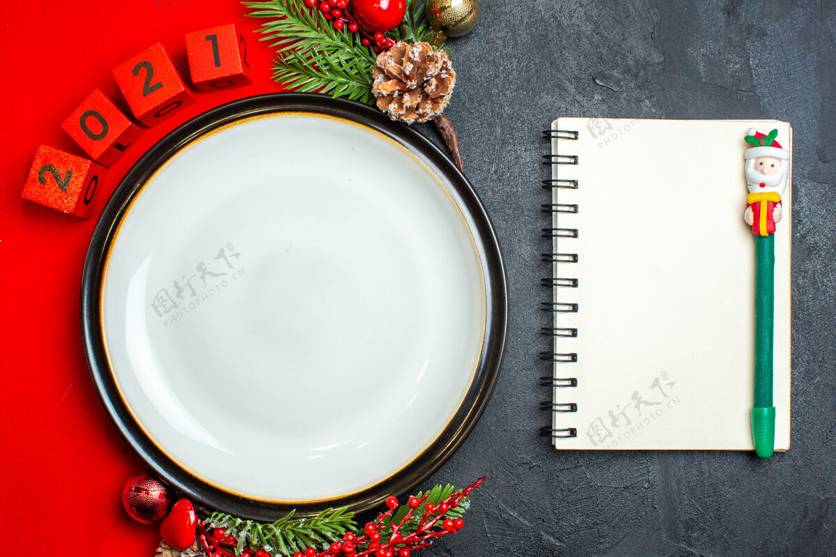 餐巾新年背景俯视图 带有餐盘装饰配件 杉木树枝和数字放在一张红色餐巾上 笔记本旁边是一张黑色桌子上的钢笔树枝餐盘冷杉