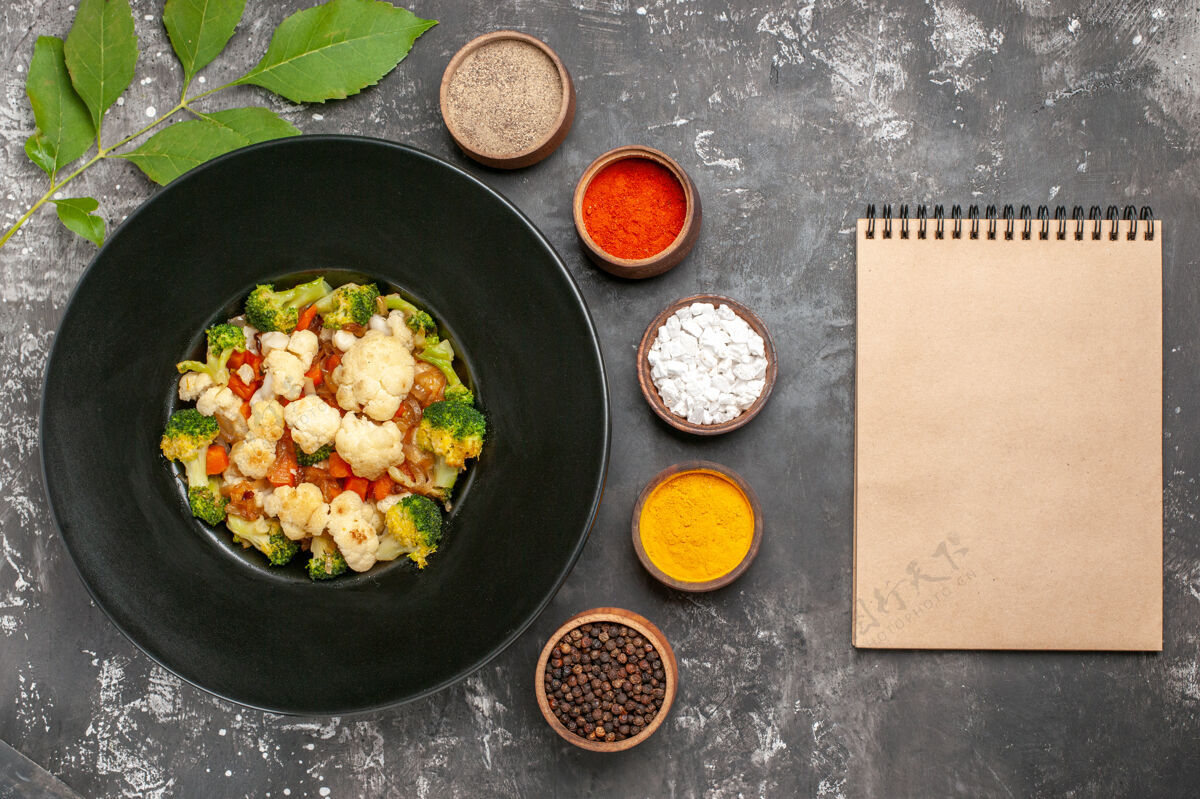 笔记本俯视图西兰花和花椰菜沙拉在黑色椭圆形盘子里不同的香料在小碗里一个笔记本在黑暗的表面上午餐厨房用具炊具
