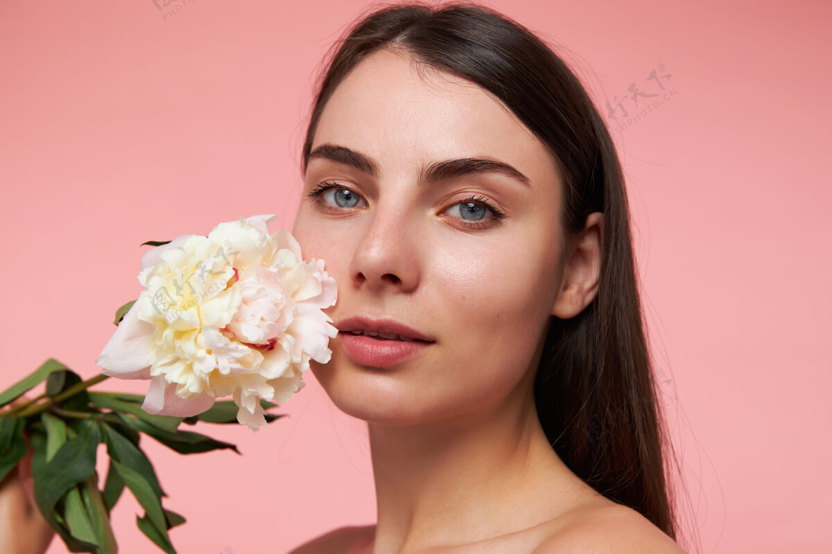 年轻年轻的女士 漂亮迷人的女人 深色长发 健康的皮肤 用一朵花抚摸脸颊观看 特写 隔离在粉彩粉色的墙上面部模特年轻