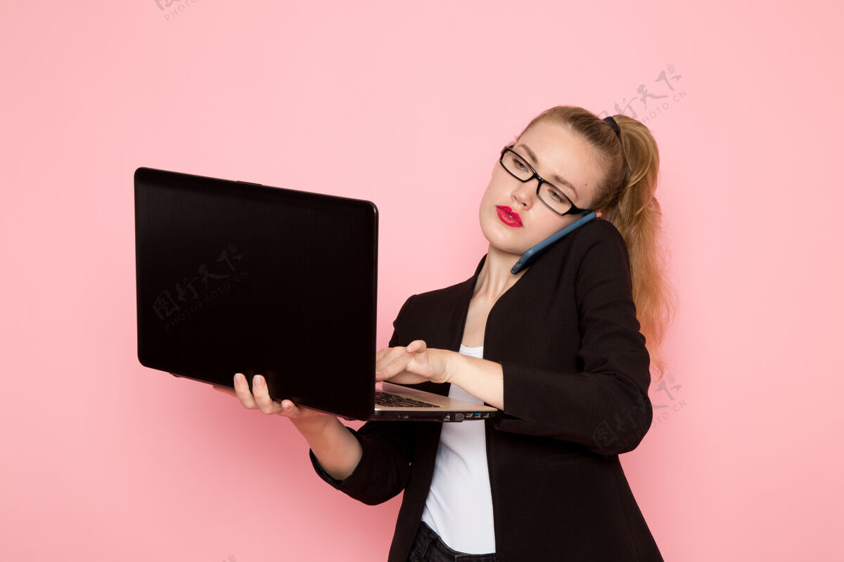 严格身穿黑色夹克的女上班族在粉色墙上打电话和使用笔记本电脑的正面图女商务会说话