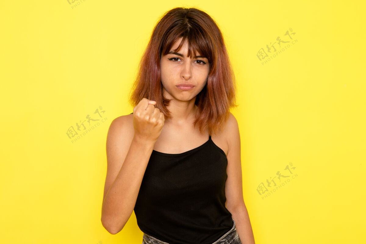 漂亮身穿黑色衬衫和灰色牛仔裤的年轻女性的正面视图 她的拳头威胁着黄色的墙壁牛仔裤女性前面