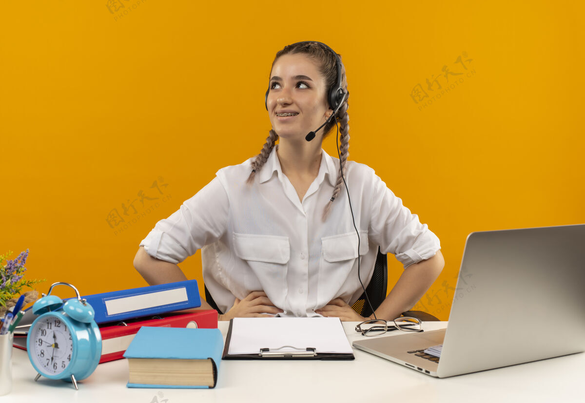 手势一个年轻的女学生扎着辫子 穿着白衬衫 戴着耳机 带着麦克风 面带微笑 坐在桌子旁 拿着笔记本文件夹 剪贴板和书放在橙色的墙上公民微笑麦克风