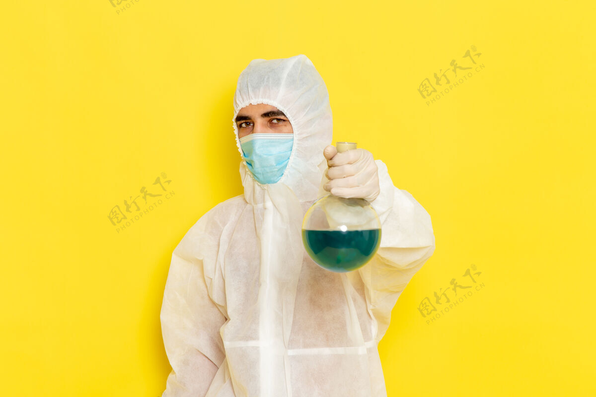 危险身穿特殊防护服的男性科学工作者的正面图 黄色墙上有带面罩的保温瓶防护健康医疗