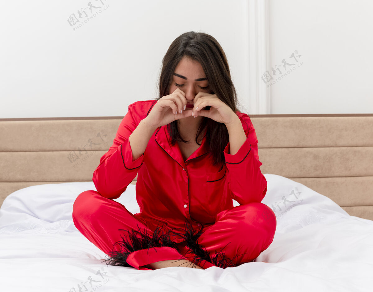 哭着穿着红色睡衣坐在床上哭泣的年轻漂亮女人在卧室里的灯光背景下哭泣睡衣年轻坐着
