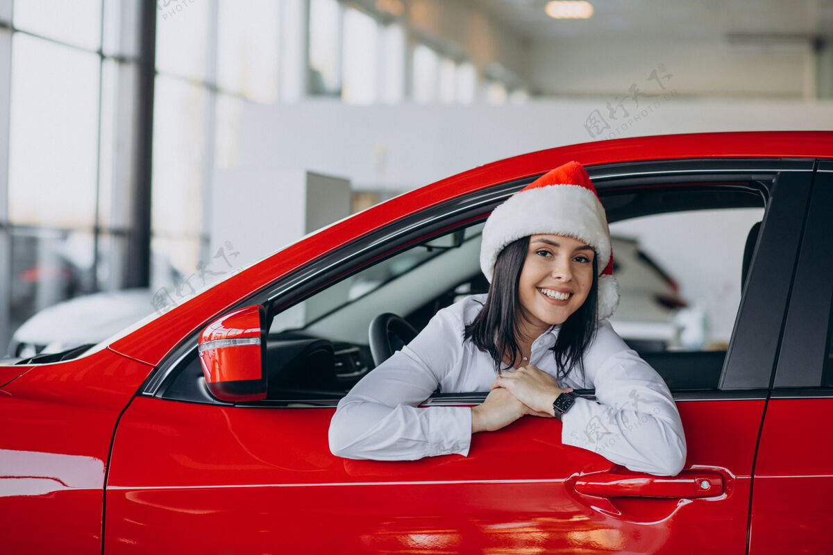 快乐一个戴着圣诞帽的女人坐在汽车陈列室里的红色汽车旁边汽车节日驾驶