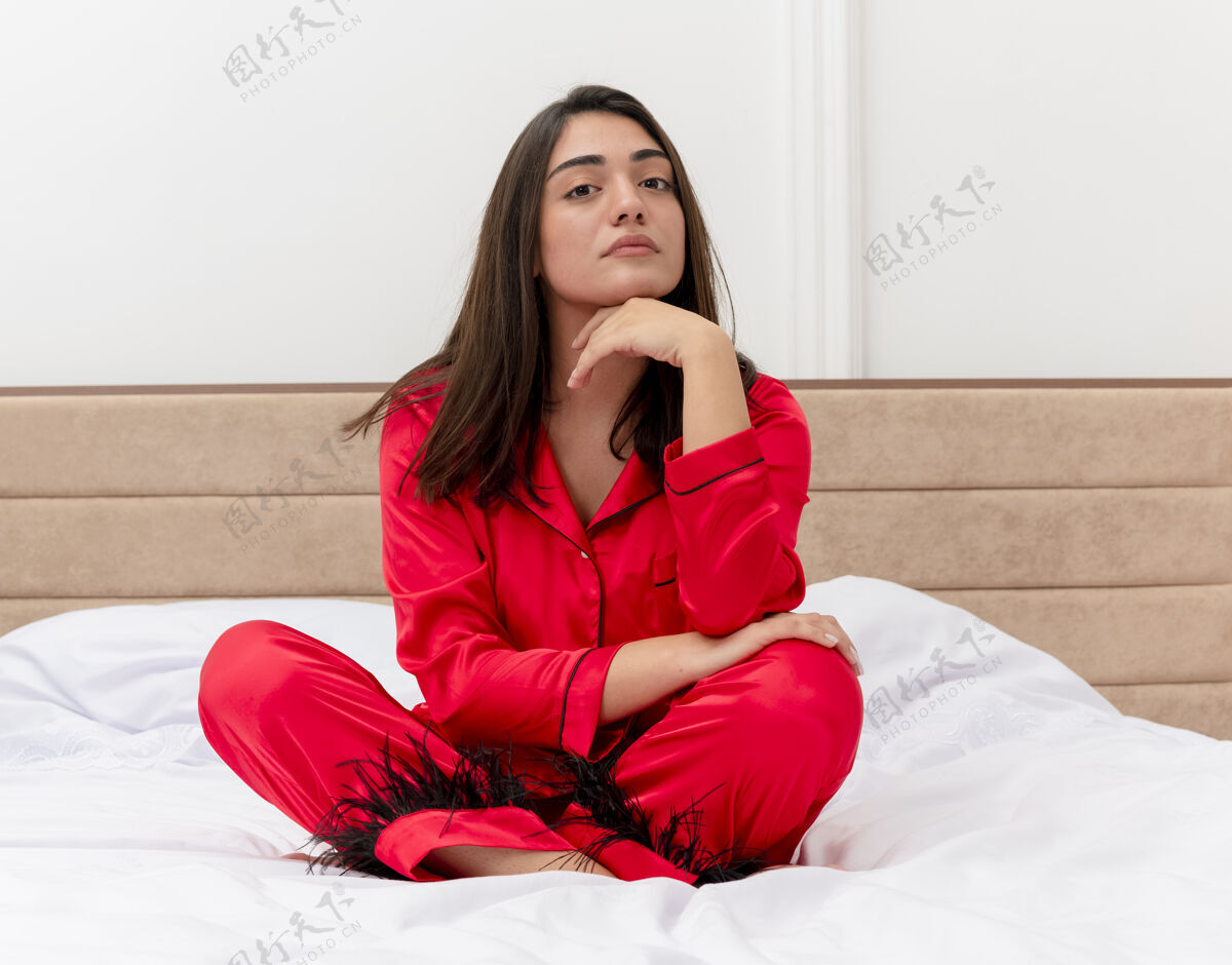 卧室穿着红色睡衣的年轻漂亮女人坐在床上 在灯光背景下严肃地看着卧室内部的摄像头漂亮脸睡衣