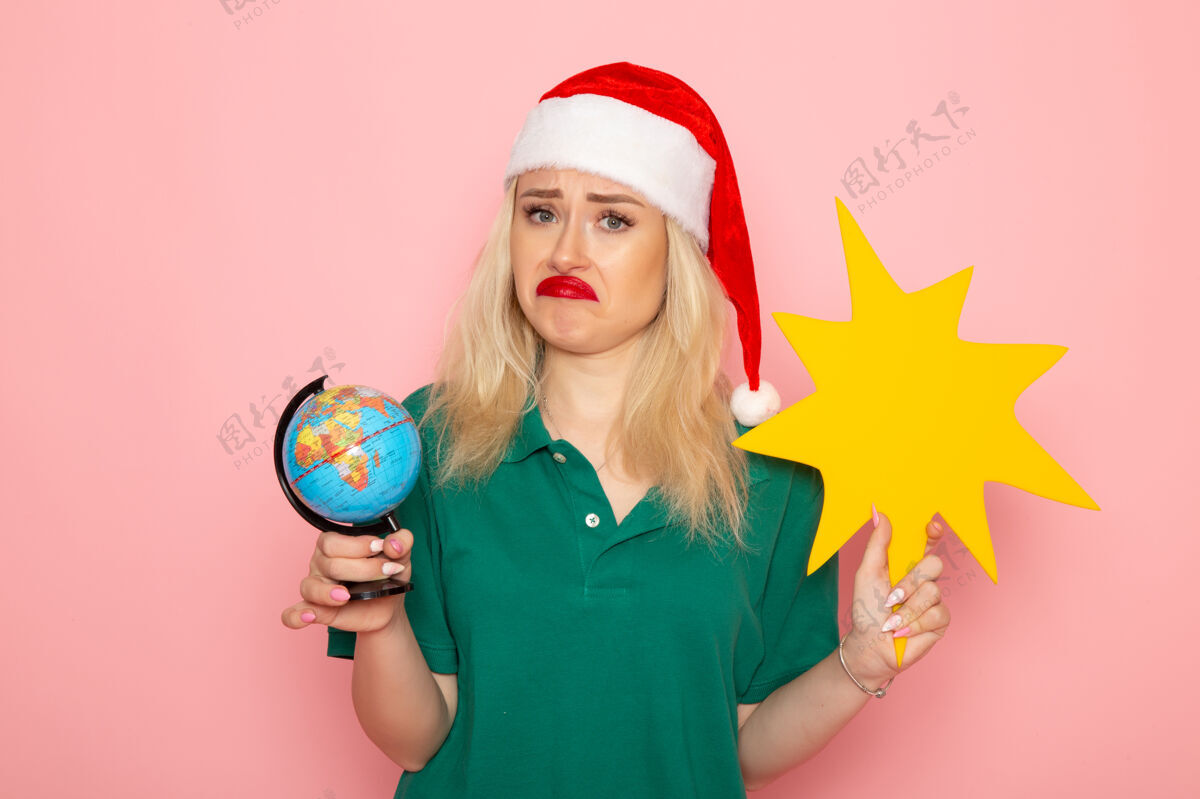 年轻的女性正面图年轻女性手持地球仪和黄色人影在粉色墙壁上的模特女假日身材圣诞节微笑