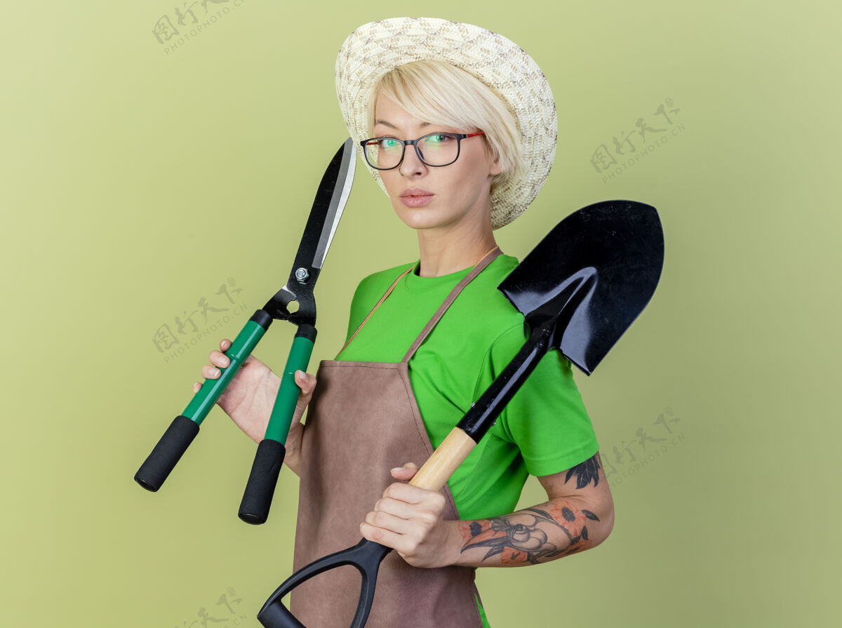 头发一位年轻的园丁 围裙上留着短发 戴着帽子 手里拿着园艺设备 站在明亮的背景下 神情严肃地看着摄像机园艺相机举行