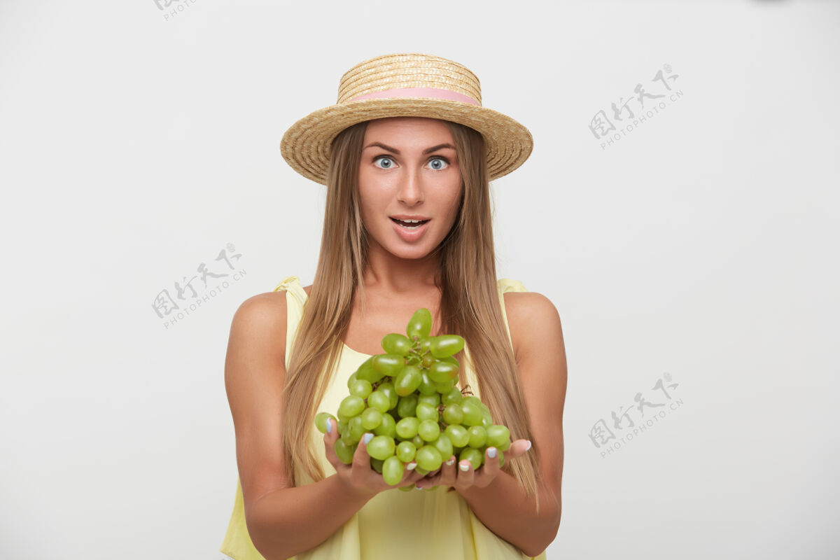 女士兴奋的年轻漂亮的长发金发女性 戴着草帽 睁大眼睛看着镜头 举着葡萄 孤立在白色背景下开放金发年轻