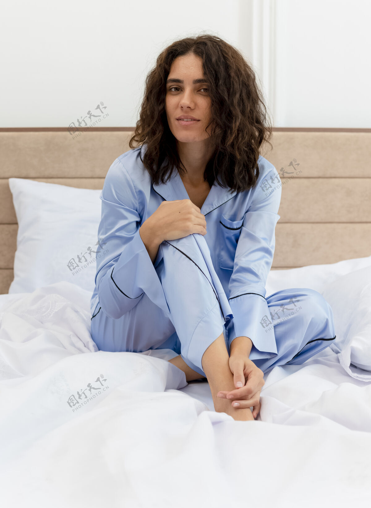 床穿着蓝色睡衣的年轻漂亮女人坐在床上看着相机 脸上带着微笑 在卧室的室内灯光背景下相机室内年轻