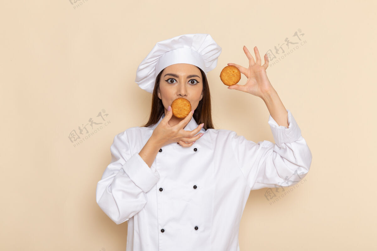 成人身穿白色厨师服的年轻女厨师正拿着小饼干站在浅白的墙上小工人饼干