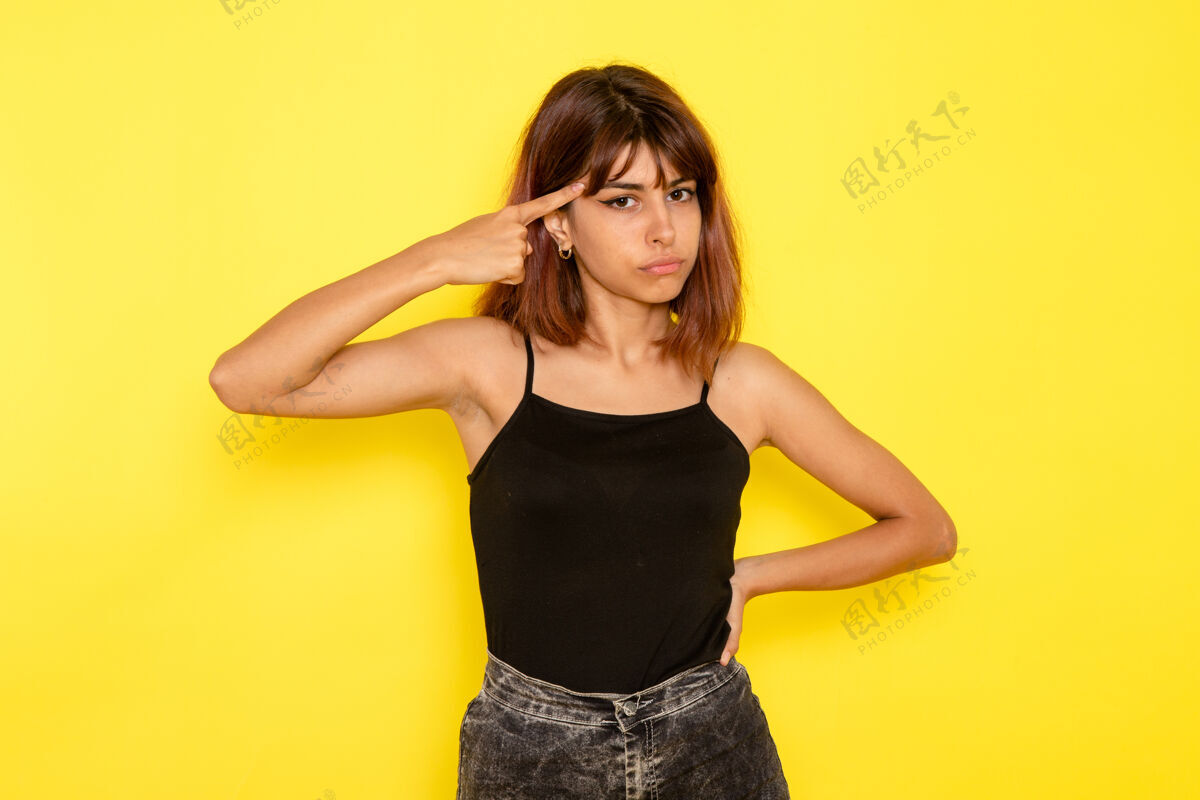 时尚身穿黑色衬衫和灰色牛仔裤的年轻女性在黄色墙壁上摆姿势的前视图情感漂亮牛仔裤