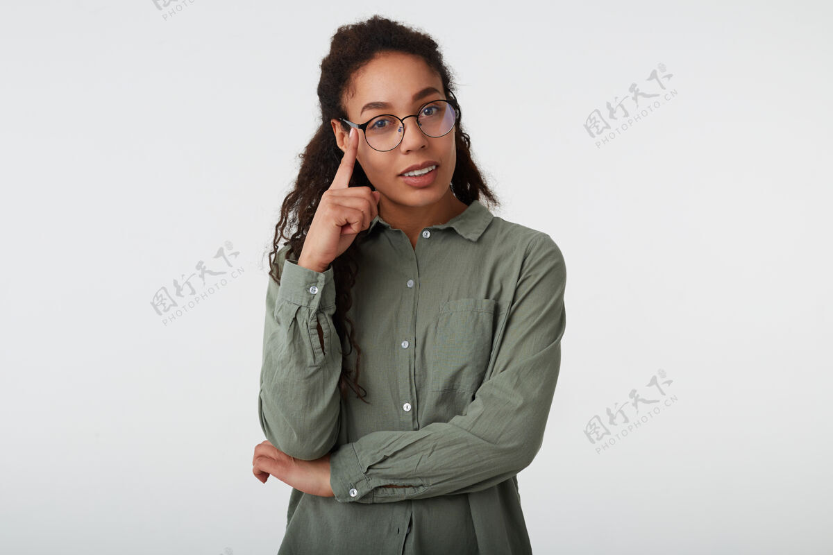 女士室内拍摄的积极年轻的棕色头发卷曲的女性 皮肤黝黑 保持抬起的手在她的眼镜 而在白色的背景下 在绿色衬衫摆姿势姿势高兴眼镜