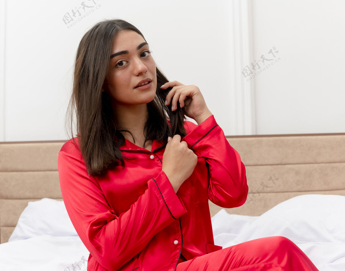 坐着穿着红色睡衣的年轻美女坐在床上讲着手机 在灯光的背景下 卧室里看起来很自信红色床说话