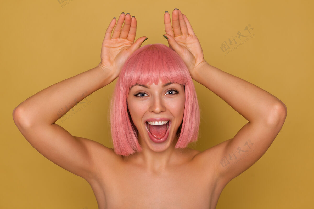 女士年轻激动的美丽粉红头发女性的肖像画 自然化妆 模仿兔子耳朵 举着手掌 愉快地看着相机 带着灿烂的微笑 在芥末背景下孤立休闲耳朵眼睛