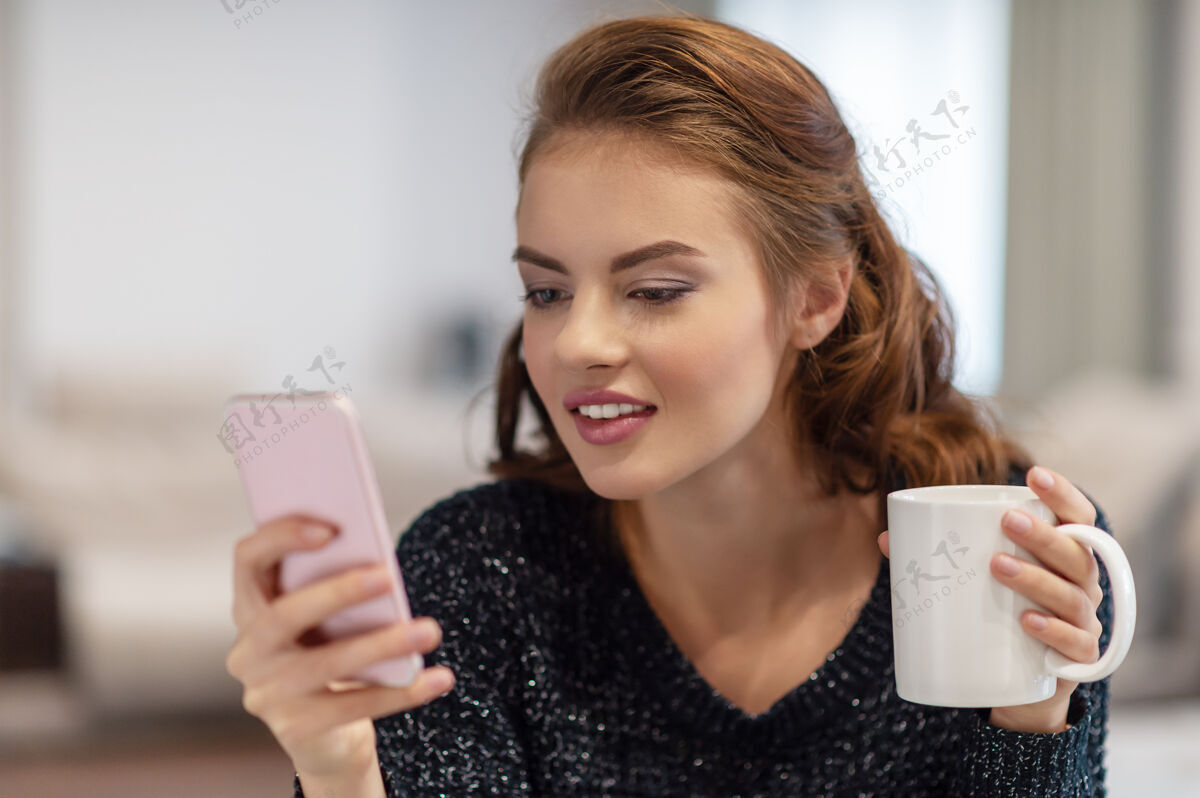 微笑漂亮的年轻女子拿着一杯咖啡看着她的智能手机女子在智能手机上输入信息迷人房间杯子
