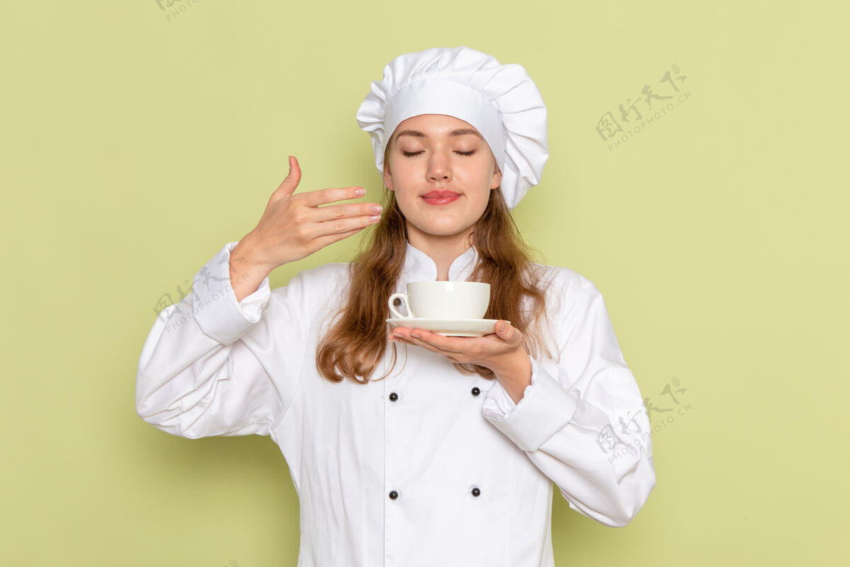 人身着白色厨师服的女厨师正拿着一杯咖啡 闻着绿墙上的味道杯子肖像烹饪