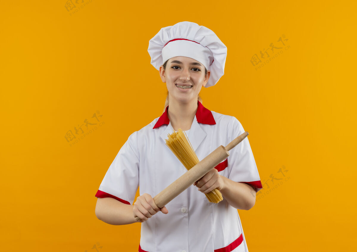 微笑微笑着的年轻漂亮厨师身着厨师制服 戴着牙套 拿着意大利面和擀面杖孤立在橙色的空间里意大利面厨师年轻