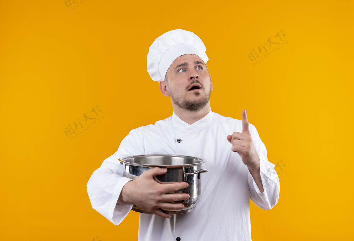 提高惊讶的年轻帅哥厨师穿着厨师制服拿着锅炉 抬起手指看着右侧孤立的橙色空间手指厨师厨师