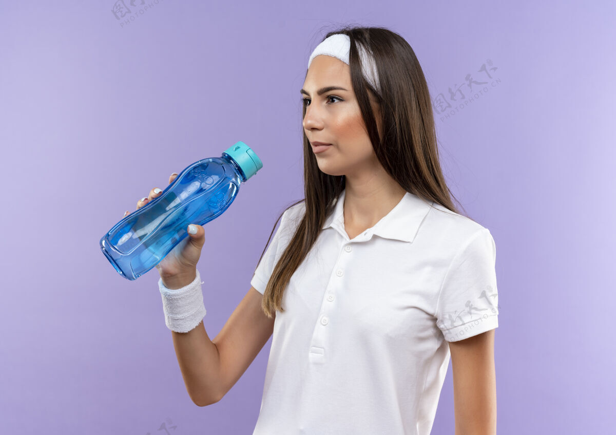看漂亮的运动女孩戴着头带和腕带拿着水瓶看着紫色空间上孤立的一面运动穿女孩