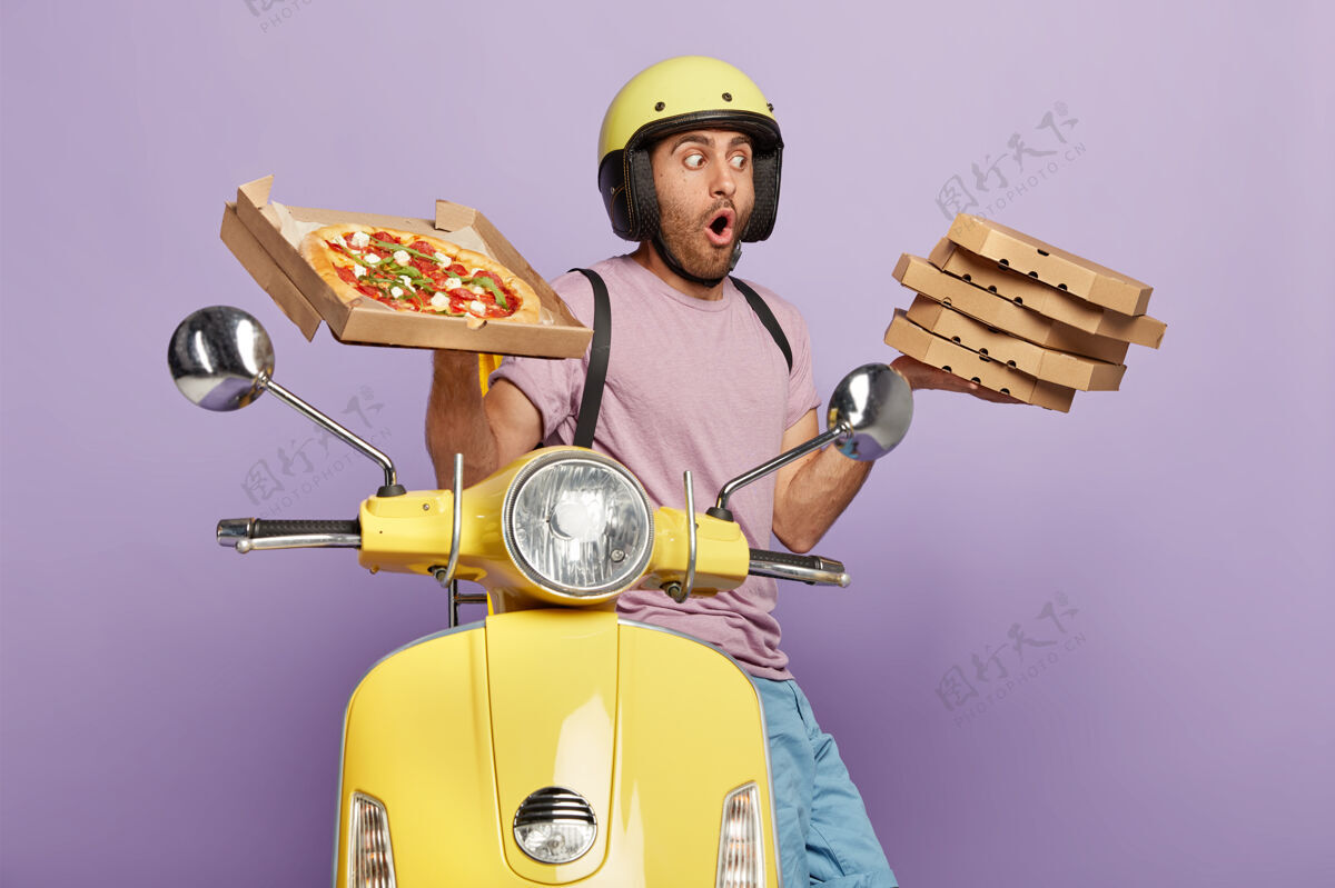 快餐震惊的送货员端着一堆美味的意大利披萨 戴着头盔 穿着休闲服 开着摩托车 运送快餐当晚餐 隔着紫色的墙美味的小吃纸箱人震惊