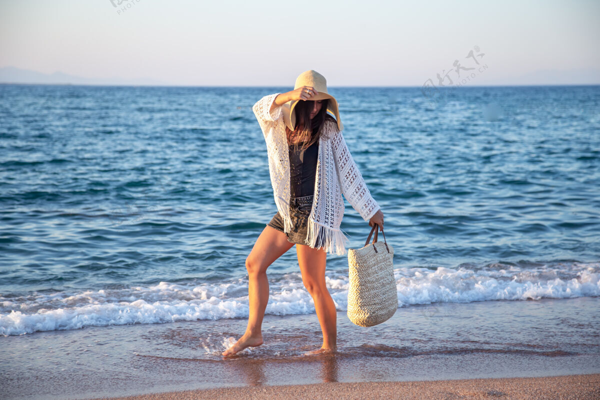 女人一个戴着大帽子 挎着柳条袋的女孩走在海边暑假的概念海岸波西米亚海滩