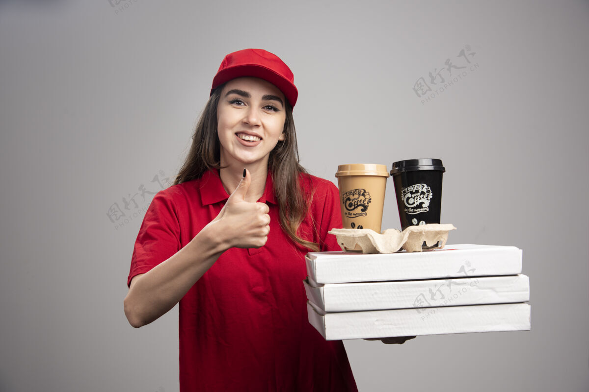 披萨快乐的送货员拿着披萨和咖啡杯在灰色的墙上竖起大拇指工人职业女性