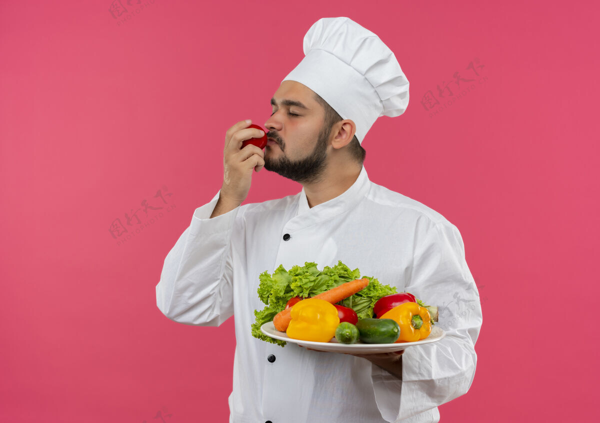 男性年轻的男厨师穿着厨师制服 拿着一盘蔬菜 闭着眼睛嗅着西红柿 被隔离在粉红色的空间里粉红年轻持有