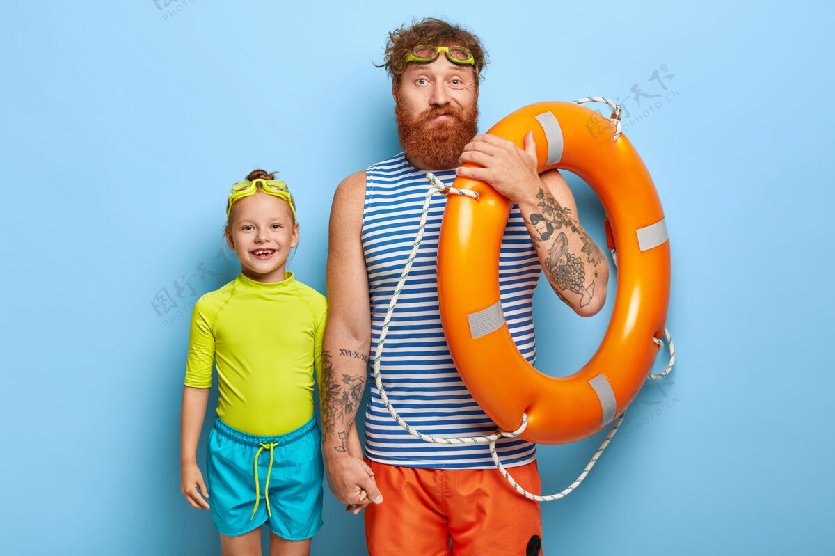 微笑家庭娱乐胡子姜爸爸牵着小女儿的手 穿着夏装 拿着游泳器材 在海边度暑假 孤零零的在蓝色的墙上 像这个季节团结女孩爸爸