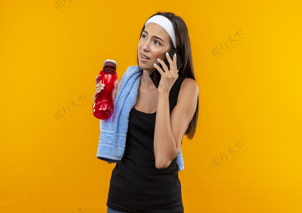 看体贴的年轻漂亮的运动女孩戴着头带和腕带打电话 看着一边拿着水瓶 肩上放着毛巾隔离在橙色的空间里橙色腕带女孩