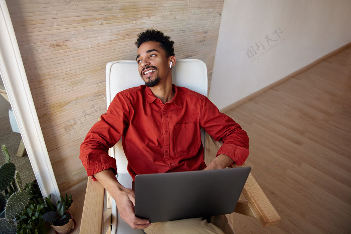 工作积极的年轻黑发胡子黑皮肤的家伙在红色衬衫后仰着他的头 而坐在椅子上 愉快地看着窗外 孤立在家里的内部年轻笔记本电脑米色
