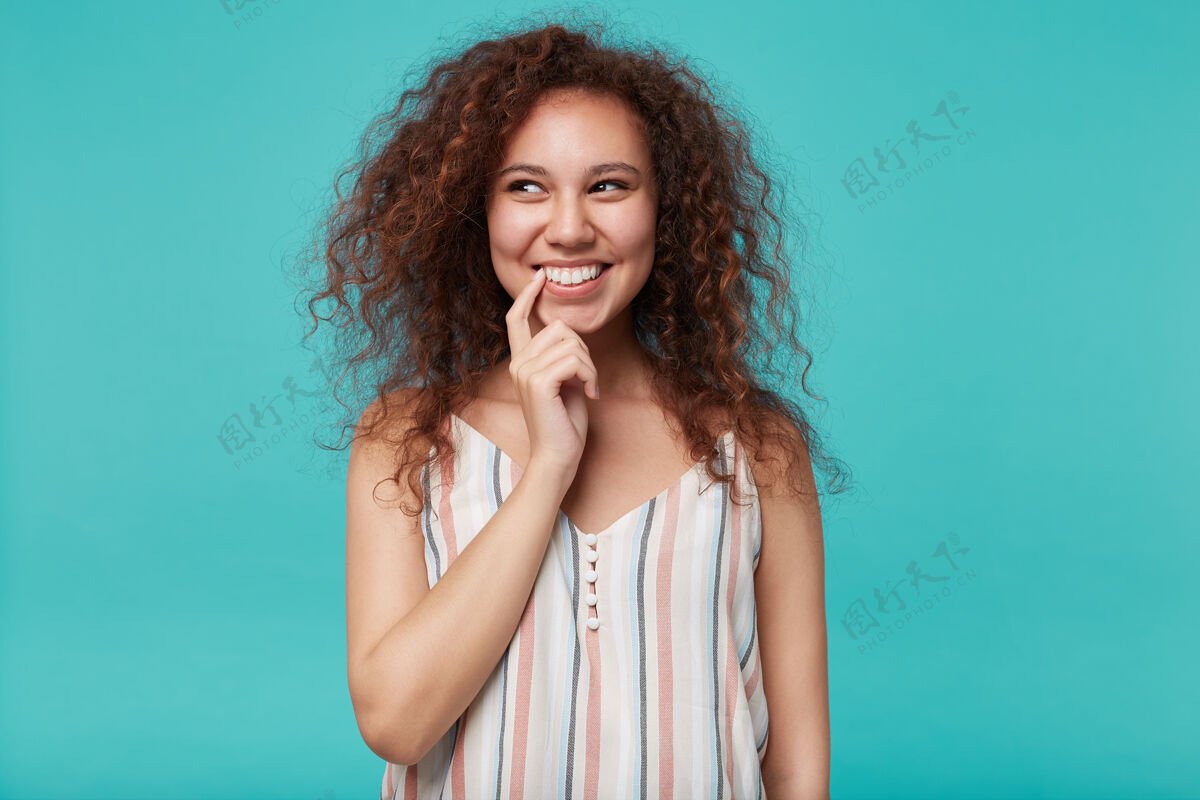 女士室内肖像 年轻漂亮的棕色卷发女士 自然妆容 微笑愉快 一边梦幻般地看着一边 食指放在嘴上 与蓝色隔离发型表情黑发